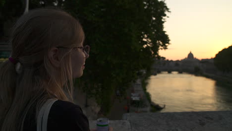Woman-sips-drink-while-looking-over-river-in-Rome-city-at-dusk,-slow-motion