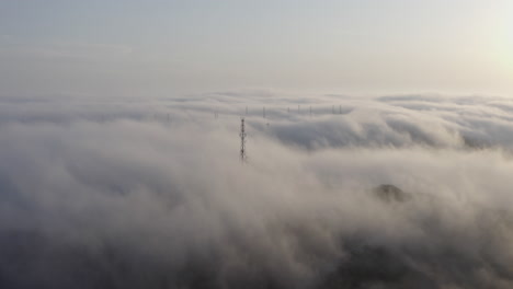 Array-of-telecommunication-towers-sticking-out-of-a-dense-heavy-fog,-moving-over-hills-in-the-afternoon