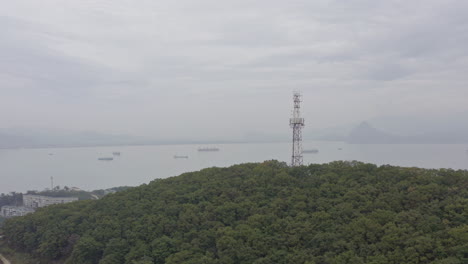 Radio-Telecom-tower-on-top-of-a-green-forest-mountain-with-the-reveal-of-bay-area-with-ships-at-anchorage-and-mountain-ridge-in-far-distance