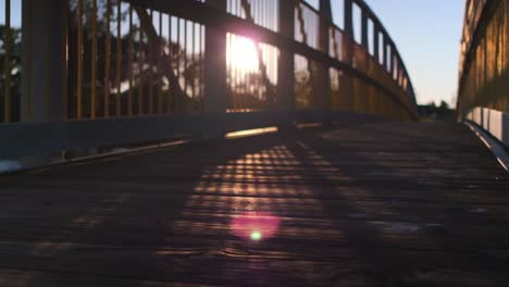 Sonne,-Die-Während-Des-Sonnenuntergangs-Durch-Die-Metallschienen-Einer-Fußgängerbrücke-über-Einen-Fluss-Scheint