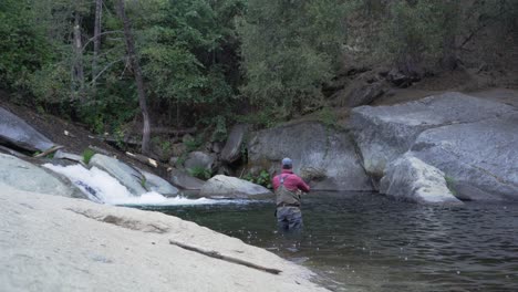 Static-shot,-man-fly-fishing-in-mountain-river-by-waterfall,-casting