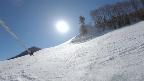 Un-Esquiador-Bajando-Una-Pendiente-Empinada,-En-El-Fondo-Un-Cielo-Azul-Con-Sol-Brillante-Y-Nieve-Saliendo-De-Los-Esquís-Vista-Desde-La-Espalda-Del-Esquiador