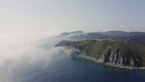 Bird's-eye-view-of-a-breathtaking-steep-rocky-coast-line-with-heavy-dense-fog-approaching-from-the-sea-and-moving-over-hill,-Far-east,-Russia,-Sea-of-Japan