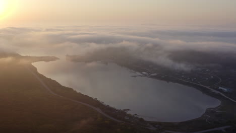 Fly-over-a-bay-with-still-water,-the-sun-reflecting-on-water-surface-and-a-dense-cloud-of-fog-coming-from-over-the-hills,-on-the-sunset