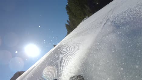 Un-Esquiador-Bajando-Una-Pendiente-Empinada,-En-El-Fondo-Un-Cielo-Azul-Con-Sol-Brillante-Y-Nieve-Saliendo-De-Los-Esquís-Vista-Desde-La-Espalda-Del-Esquiador