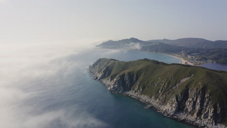 Bird's-eye-view-of-a-breathtaking-steep-rocky-coast-line-with-heavy-dense-fog-approaching-from-the-sea-and-moving-over-hill,-Far-east,-Russia,-Sea-of-Japan