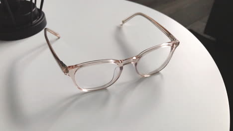 Modern-eyeglasses-in-light-colour-lie-on-a-white-table,-bright-glasses-frame,-slow-motion-footage