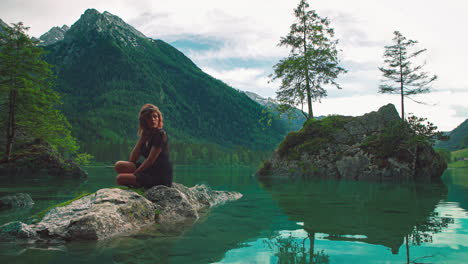 In-this-4k-UHD-Cinemagraph-a-young-brunette-woman-is-sitting-on-a-rock-at-the-scenic-mountain-lake-Hintersee-in-Bavaria,-a-perfect-travel-destination-in-Germany-with-the-water-moving-gently