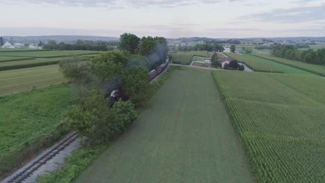 Vista-Aérea-De-Un-Tren-De-Pasajeros-De-Vapor-Que-Se-Aproxima-En-La-Campiña-Amish-En-Un-Día-De-Verano