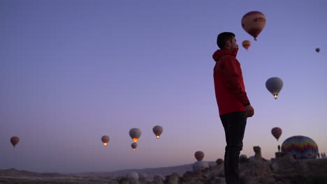 Slow-motion-rotating-view-of-a-man-surrounded-by-rising-hot-air-balloons-at-dawn-in-Cappadocia,-Turkey