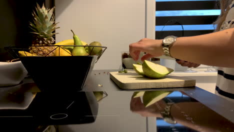 Slowmotion:-Young-woman-in-the-kitchen-preparing-a-fresh-dessert-with-pear,-girl-slicing-a-green-pear-on-a-wooden-board