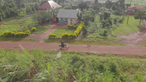 Luftaufnahmeverfolgung-Von-Der-Seite-Eines-Afrikanischen-Mannes,-Der-Einen-Helm-Auf-Einem-Motorrad-Trägt,-Das-Auf-Einer-Unbefestigten-Straße-Durch-Ein-Ländliches-Dorf-Fährt