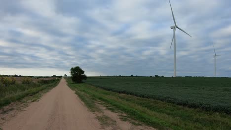 En-Un-Camino-De-Ripio-Rural-Viendo-Turbinas-Eólicas-Giratorias-Que-Se-Encuentran-En-Un-Campo-De-Soja-A-Lo-Largo-De-Un-Camino-De-Ripio-En-La-Zona-Rural-De-Nebraska,-EE.UU.-En-Un-Día-Nublado