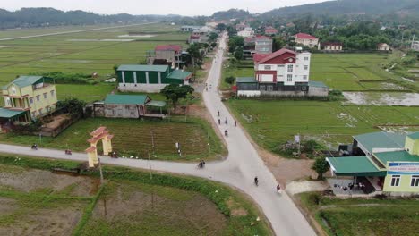 Luftaufnahme-Von-Vietnamesen,-Die-Motorräder-Auf-Der-Fahrbahn-Neben-Der-Reisfeldfarm-Und-Den-Häusern-Fahren