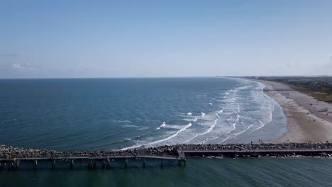 Sunny-coastline-of-Port-Canaveral,-Florida-with-waves-rolling-onto-the-sandy-beach,-flat-sea-and-clear-blue-sky