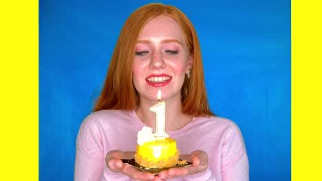 A-young-woman-stands-smiling-in-a-bright-studio-against-a-blue-and-yellow-backdrop,-and-holds-a-small-cake-with-a-number-one-shaped-candle