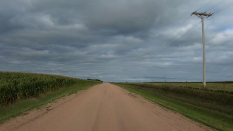 POV-while-driving-on-a-rural-gravel-road-with-small-seed-business,-fields,-power-lines,-telephone-lines-and-farm-houses-along-the-road