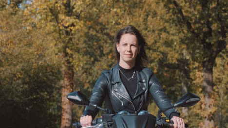 Moving-view-of-pretty-smiling-European-young-woman-driving-a-motorbike-wearing-leather-jacket-in-forest-with-vibrant,-colorful-golden-autumn-leaves-on-sunny-day