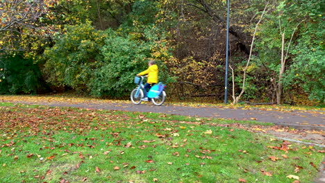 People-are-riding-their-bikes,-View-of-the-bike-path-in-the-autumn-season,-the-trees-have-colourful-leaves,-falling-leaves-on-the-road-and-green-grass