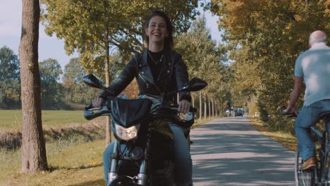 Front-view-of-beautiful-smiling-European-Female-motor-biker-in-leather-jacket-driving-On-road-passing-cyclists-with-autumn-leaf-color-Trees-in-forest-on-sunny-day