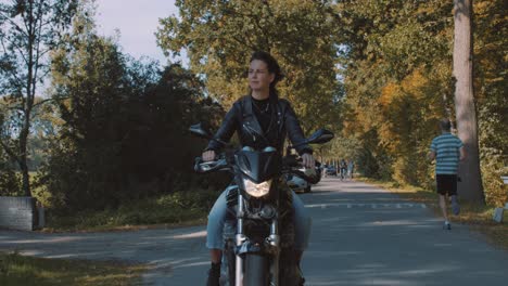 Pretty-smiling-European-young-woman-driving-a-motorbike-wearing-leather-jacket-in-forest-with-vibrant,-colorful-golden-autumn-leaves-on-sunny-day