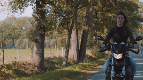 Pan-from-forest-fields,-farmland-to-Front-view-of-beautiful-European-Female-Biker-wearing-leather-jacket-On-road-with-autumn-leaf-colored-trees-on-sunny-day