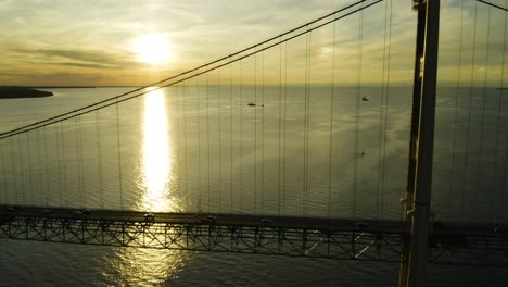 Aerial,-traffic-on-large-suspension-bridge-with-sunset-over-ocean-in-background