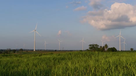 Wind-Turbines-shooting-out-of-a-Farmland-while-the-Sun-is-setting-as-Trucks-Pass-by-making-Clouds-of-Dust,-clean-alternative-energy-in-Thailand-and-mainland-Southeast-Asia