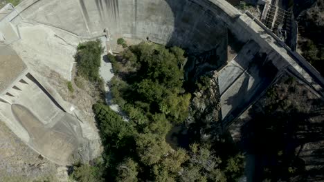 Aerial-shot-looking-straight-down-flying-over-large-curved-concrete-dam,-glides-over-dark-glassy-reservoir-and-small-moving-boat-beyond-dam