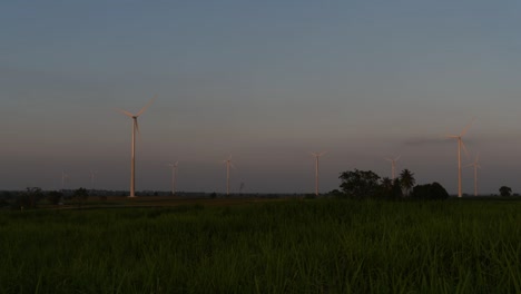 Wind-Turbines-shooting-out-of-a-Farmland-while-the-Sun-is-setting-as-Trucks-Pass-by-making-Clouds-of-Dust,-clean-alternative-energy-in-Thailand-and-mainland-Southeast-Asia