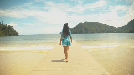 A-girl-in-a-short-dress-walks-out-from-a-cabana-down-the-jetty-with-waves-crashing-at-the-shoreline-with-mountains-in-the-background