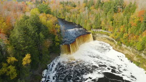 Aerial-backwards-tilt-up,-wide-brown-river-waterfall-in-autumn-forest