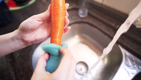 Frauenhände-Waschen-Einige-Karotten-Unter-Wasser-Aus-Einem-Küchenhahn-Mit-Einem-Schwamm-In-Zeitlupe