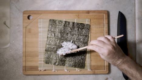 Top-down-time-lapse-of-two-hands-with-wooden-spoon-and-cutting-tablet-on-kitchen-counter-spreading-rice-with-chai-seeds-on-a-mat-and-seaweed-Nori-sheet-to-make-Sushi