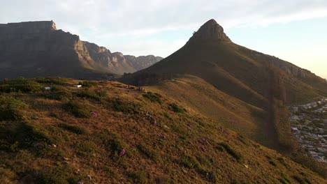 Dramatic-Pull-Away-Cinematic-Aerial-Shot-of-Lion's-Head-Peak-with-Table-Mountain-and-Signal-Hill-During-Golden-Hour-Sunset