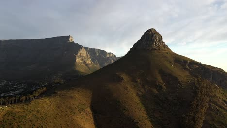 Cinematic-Aerial-Rotational-Shot-of-Cape-Town's-Lion's-Head-Peak-with-Table-Mountain-and-Signal-Hill-During-Golden-Hour-Sunset