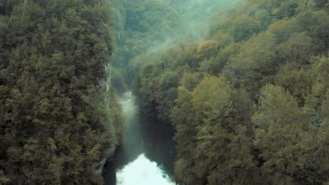 Slow-aerial-shot-drifting-lazily-down-a-misty-tree-lined-river,-with-a-rock-cliff-on-left-side