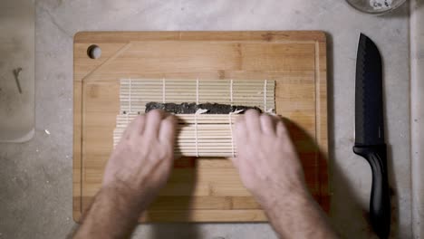 Time-lapse-of-top-down-view-on-two-hands-on-wooden-cutting-tablet-on-kitchen-counter-rolling-ingredients-in-a-mat-and-seaweed-Nori-sheet-making-a-Sushi-roll