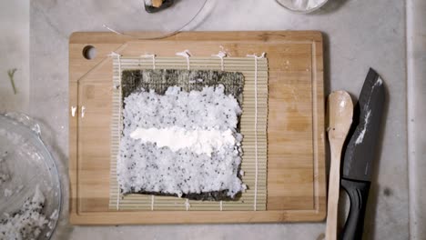 Time-lapse-of-top-down-view-on-two-hands-on-wooden-cutting-tablet-on-kitchen-counter-spreading-rice-with-chai-seeds-on-a-mat-and-seaweed-Nori-sheet-to-make-Sushi