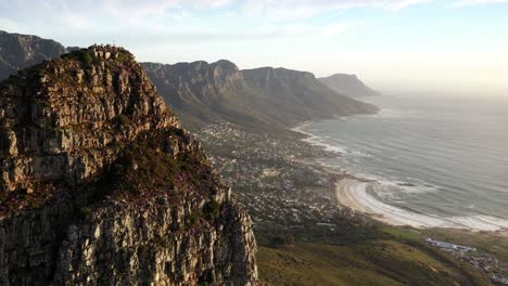 Fly-By-Cinematic-Aerial-Shot-of-Cape-Town's-Lions-Head-Peak-Revealing-Camps-Bay-and-Table-Mountain-National-Park-During-Golden-Hour-Sunset