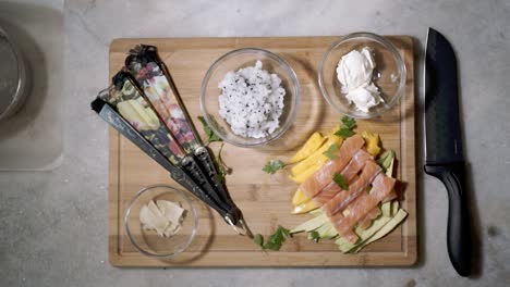Exhibición-De-Ingredientes-Para-Sushi-Con-Hierbas-Verdes-Frescas-Que-Caen-Vistas-Desde-Arriba-Hacia-Abajo-En-El-Mostrador-De-La-Cocina-Con-Tazones-De-Ingredientes-Que-Incluyen-Arroz,-Salmón-Y-Frutas-Listos-Para-Hacer-Sushi
