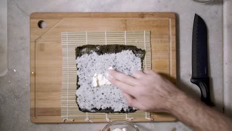 Top-down-view-of-two-hands-on-wooden-cutting-tablet-on-kitchen-counter-spreading-cream-with-fingers-on-the-chai-seed-rice-on-a-mat-and-seaweed-Nori-sheet-to-make-Sushi