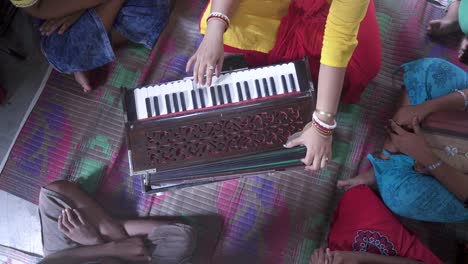 Married-Indian-teacher-teaches-music-with-harmonium-at-rural-classroom-with-students-gathered-around,-unrecognizable,-top-down-shot