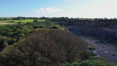 aerial-view-of-tree-tops-to-the-side-of-abandoned-quarry-with-blue-sky-and-green-fields-in-the-distance