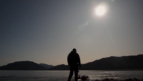 Silhouette-Eines-Mannes-Zu-Fuß-Mit-Blick-Auf-Die-Sonne-Und-Die-Berge-Auf-Dem-Schimmernden-See
