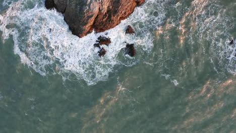 Birds-Eye-Aerial-Shot-of-Waves-Crashing-against-a-Rocky-Outcrop-with-Sea-Lions-at-Sunset