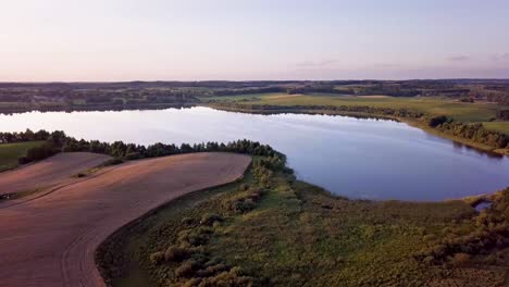 Aerial-Shot-of-a-Small,-Peaceful-Lake-Surrounded-by-Trees-and-Farms-at-Sunset