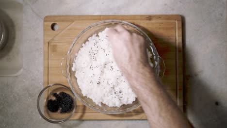 Blick-Von-Oben-Nach-Unten-Auf-Die-Küchentheke-Mit-Einer-Glasschale-Mit-Sushi-Reis-Und-Einer-Hand,-Die-Chai-Samen-Auf-Dem-Teller-Als-Teil-Eines-Rezepts-Teilt