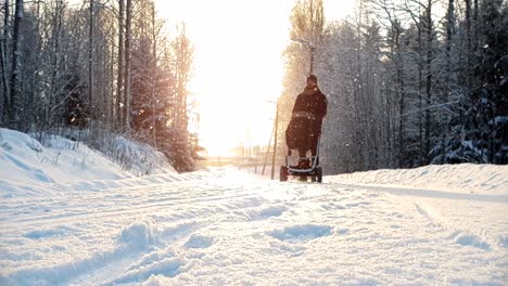 Low-angle-slow-motion-shot-of-man-walking-with-baby-wagon-in-snowy-winter-weather,-with-beautiful-golden-sunshine-in-the-background