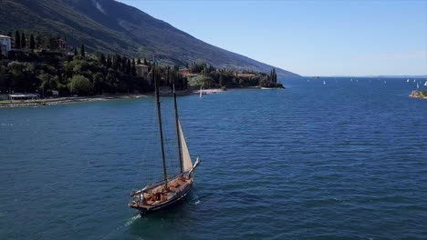 Luftaufnahme-Eines-Historischen-Segelschiffs-Auf-Dem-Lago-Di-Garda-Mit-Der-Küste-Im-Hintergrund-An-Einem-Sonnigen-Tag